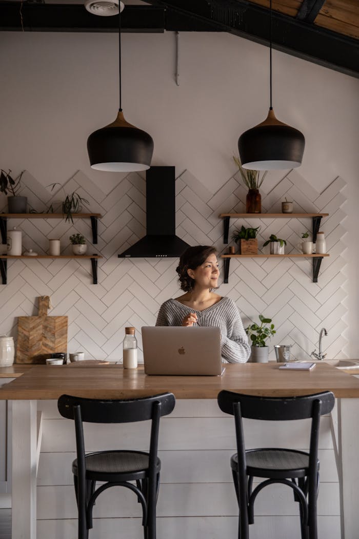 project-02-c Cheerful female in gray sweater sitting at wooden counter with laptop and bottle of milk while browsing internet on netbook during free time at home and looking away