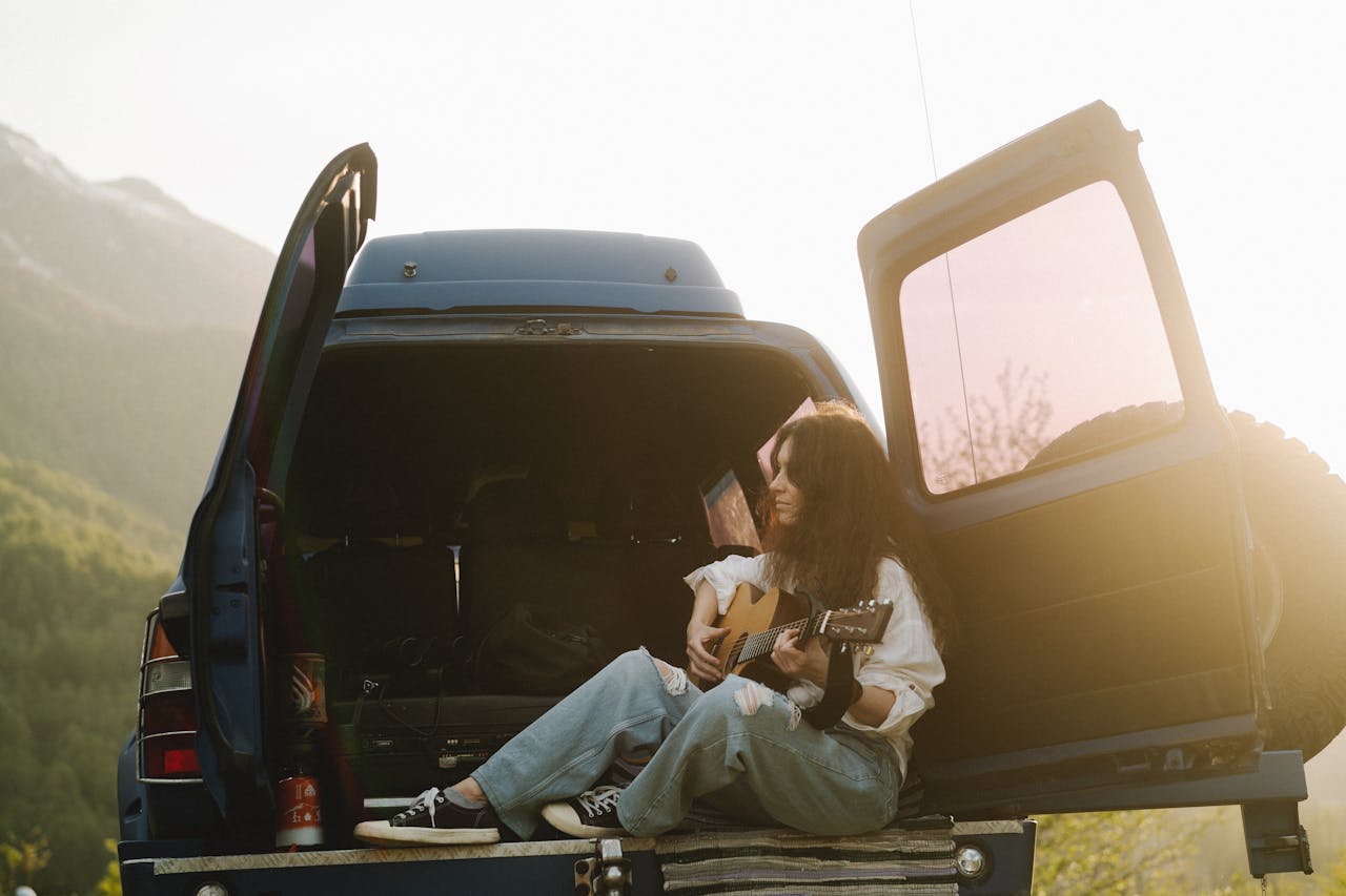 project-03-c Woman playing guitar near open van at sunset in a scenic mountain location.